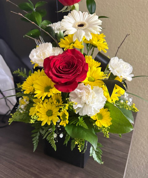 Colorful flower bouquet in a black vase with a red rose, white carnations, white gerbera daisies, and yellow daisies with greenery.