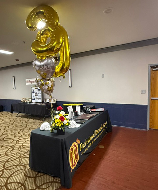 Table setup for a celebration with gold '31' balloons and a flower bouquet in front of a black draped table, event banners in the background.
