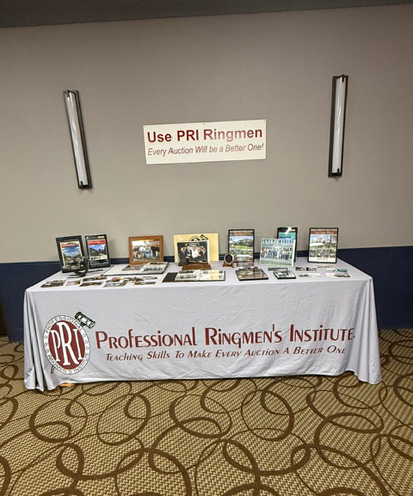 Exhibit table for the Professional Ringmen's Institute with framed photos and brochures on a white tablecloth labeled PRI logo. The backdrop includes a sign promoting PRI Ringmen and a patterned carpeted floor.