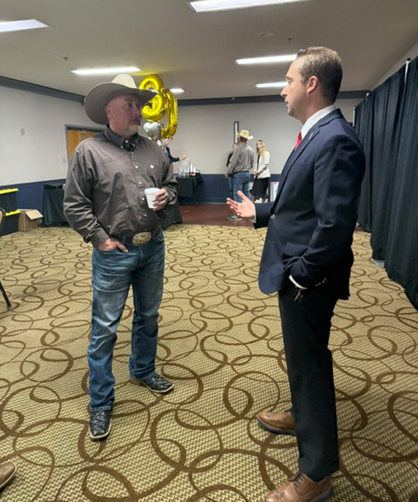 Two men chat at a social event: one in a cowboy hat and jeans with a coffee cup, the other in a navy suit and brown shoes.