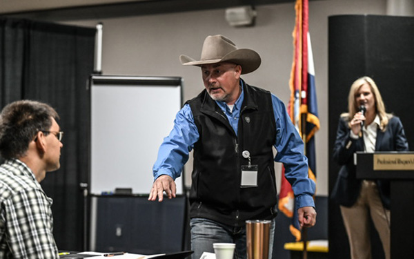 Cowboy-hatted man in a blue shirt and black vest gestures toward a seated attendee during a meeting, with a presenter at a podium in the background.