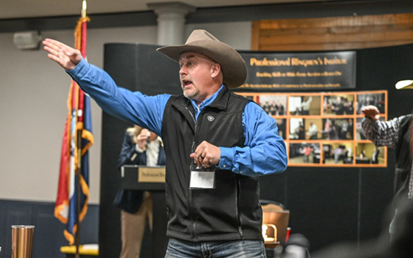 Man wearing a cowboy hat and blue shirt gestures with his left arm while speaking at a presentation in a conference room, with a collage display and flag in the background.