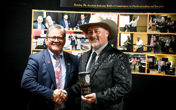 Two men in suits shake hands at an awards event; one wears a cowboy hat and holds a trophy, with a photo collage backdrop.
